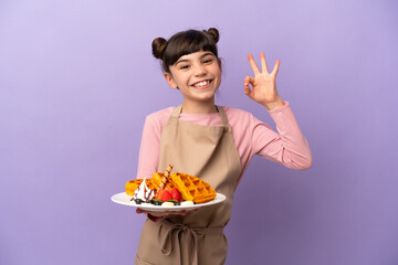 Little caucasian girl holding waffles isolated on purple background showing ok sign with fingers