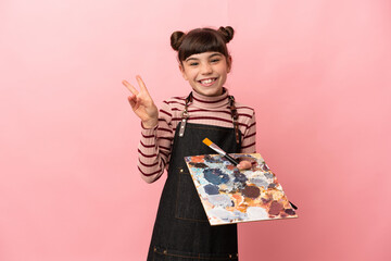 Little artist girl holding a palette isolated on pink background smiling and showing victory sign