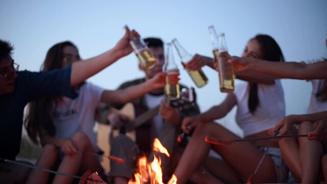 Friends sit around bonfire cheering and toasting beer bottles, playing guitar, and frying sausages on sandy beach. Young group of men and women drink beverages, singalong. Guy playing guitar in dusk.