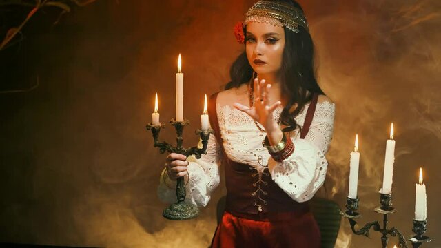 Fantasy woman gypsy in red vintage dress. Girl witch fortune teller holds candlestick burning candles. Room in smoke. Spiritual mystical seance. Witch waving hands create magic, spell, read future.