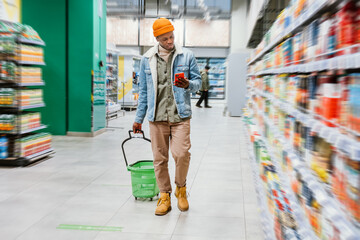 Stylish black guy with shopping basket and smartphone walks past shelves with different goods in...
