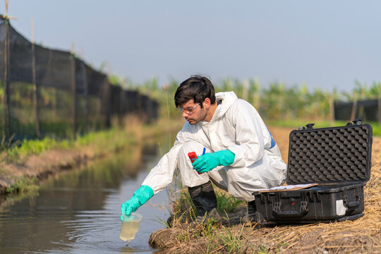 Man Technician In Full Body Protective Suit Collecting Sample Of Water .Portable Water Quality Measurement .Water Quality For Agriculture. Checking Water Ph On Field.