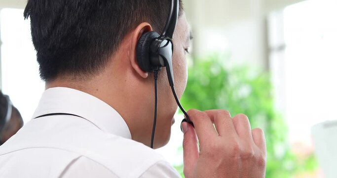 Back view of business Asian man callcenter worker in headsets working with desktop computer at telemarketing customer service agents. Close up headset.