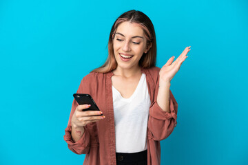 Young caucasian woman isolated on blue background looking at the camera while using the mobile with surprised expression