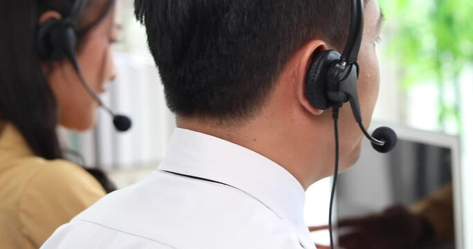 Back view of business Asian man callcenter worker in headsets working with desktop computer at telemarketing customer service agents. Close up headset.
