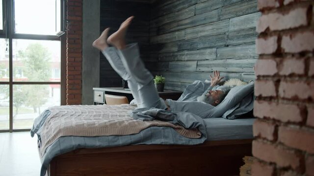 Slow motion of mature grey-haired man wearing pajamas jumping in bed at home enjoying rest alone. Relaxation and modern apartment interior concept.