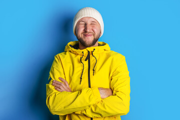 young man in a yellow jacket with closed eyes holds his arms crossed on a blue background