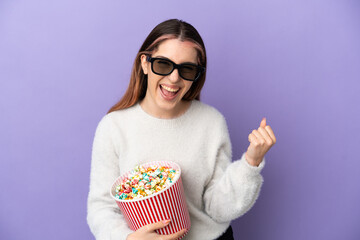 Young caucasian woman isolated on blue background with 3d glasses and holding a big bucket of popcorns