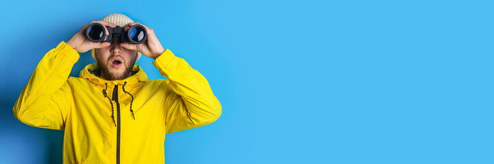 surprised young man in a yellow jacket looks through binoculars to the side on a blue background. Banner