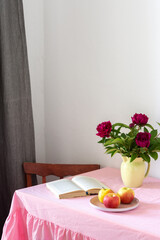 Part of the home interior, a table with a pink tablecloth with a chair near the window with a gray curtain, a beautiful bouquet of peonies stands on the table, a plate with apples and an open book
