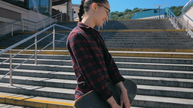Young attractive millennial woman walking along the pedestrian promenade with a skateboard in her hands. Young woman skateboarder climbs the steps turns around and smiles while looking at the camera