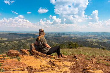 Naklejka na ściany i meble Tourist girl hiker resting at the top of the mountain and enjoying amazing landscape view of mountains and beautiful sky