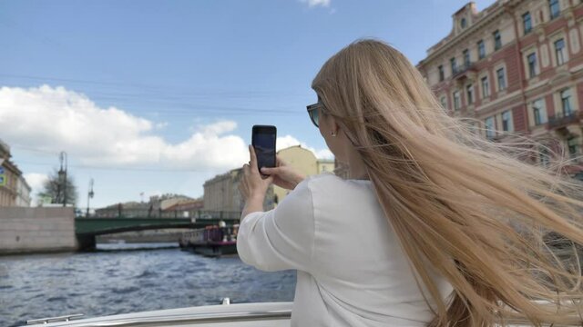Long haired blonde lady makes photo of historical bridge and river bank with buildings sailing tourist boat close backside view
