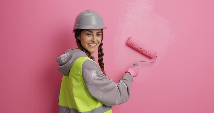 Painting service. Glad hard working professional female painter in safety clothes smiles pleasantly paints wall with pink paint uses roller does house renovation and repair has two combed pigtails.