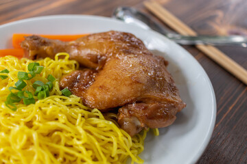 Roasted chicken thighs and drumsticks are served with yellow noodles and fresh vegetables in a white plate. Chicken thighs baked in a sweet and delicious sauce are served with hot yellow noodles.