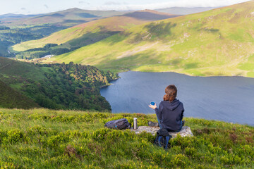 Naklejka na ściany i meble Woman having a cup of coffee and enjoying the view over Lough Tay after a hiking in Wicklow Mountains, Ireland