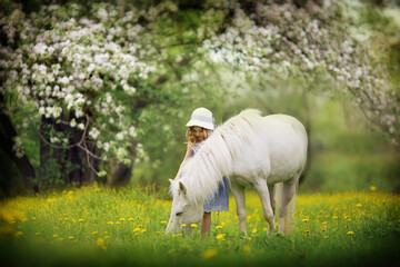 A cute little girl in a white hat caressing her little pony in the blooming garden on a sunny...