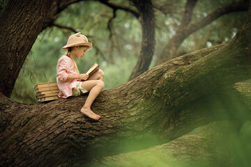 A cute little girl with blonde hair and a straw hat is reading a book in a big tree. Children and...