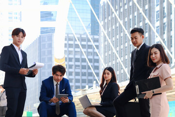 Group of five young Asian businesspeople socializing at modern city downtown with skyscraper building as blurred background., businessman and woman have meeting at outside office.