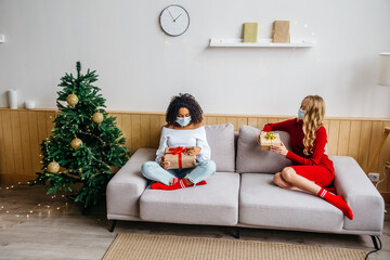 Celebrating Christmas and New Years during pandemic Covid-19 concept. Two multiethnic women in protective masks sitting on the couch and exchange gifts.