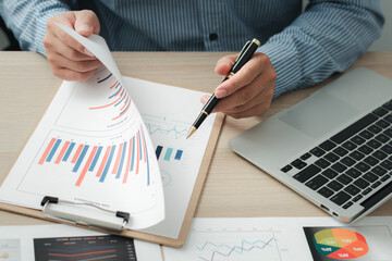 Businessman holding pen and pointing paper chart summary analyzing annual business report with using laptop at room office desk.