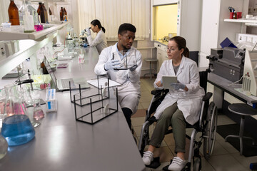 Female chemist in wheelchair using tablet and looking at mouse in glass box held by African male...