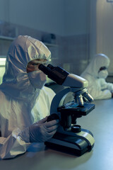 Female microbiologist in coveralls and other protective workwear looking in microscope