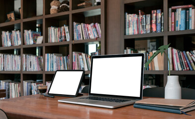 Mockup blank screen laptop computer and tablet on wooden table in library.