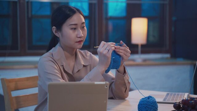 Young Adult Asian Woman Doing Hobby By Sewing And Knitting A Crochet Handmade In Living Room At Night By Learning And Study From Internet Using Laptop. New Normal Lifestyles Working At Home Concept