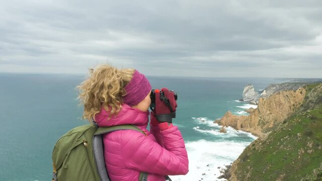 happy blonde girl tourist in pink jacket with backpack takes picture of beautiful ocean from viewing point on nasty day