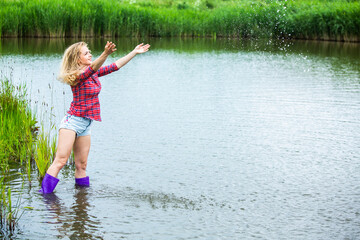 The blonde girl is having fun in the river in rubber boots in the country. Young woman on a lake enjoy the life.
