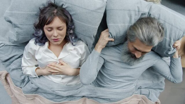 Young woman wearing pajamas is snoring in sleep while irritated husband is covering head with pillow feeling annoyed and unhappy. Sleep problems and lifestyle concept.