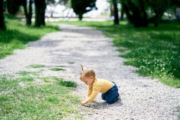 Little girl sits on her knees on a gravel path in a green park