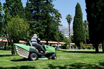 Man rides a green large lawn mower in the park - obrazy, fototapety, plakaty