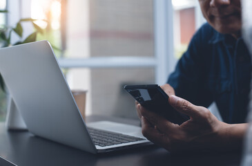 Asian casual business man using mobile phone during online working on laptop computer at home