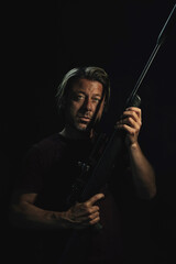 Dark portrait of blond haired man in dark brown t-shirt holding a rifle.