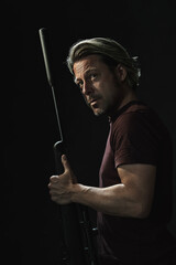 Dark portrait of blond haired man in dark brown t-shirt holding a rifle and looking over his shoulder.