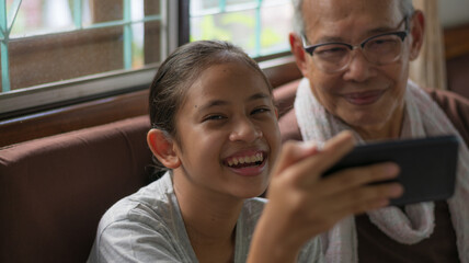 Cheerful adorable Thai girl holding mobile phone enjoy taking selfie photo with her senior grandfather in living room. Bonding relationship in family. - 440427039