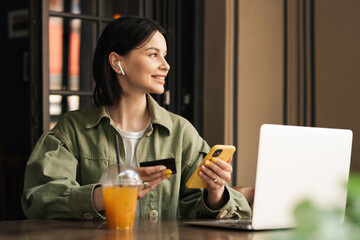 Cheerful Young Woman Holding Credit Card and Smartphone Making Online Shopping While Sitting at a Cafe Table with Laptop and Cocktail, Looking Away