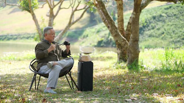 Asian senior man sit on outdoor chair using digital camera photography and taking note in the book at public park. Elderly retired male relax and enjoy outdoor leisure activity and hobby in sunny day.