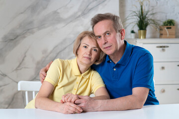 Fototapeta premium Happy beautiful elderly senior European couple in love. Attractive mature woman and handsome retired man hugging each other, sitting at table at home together, looking at camera. Retirement, happiness