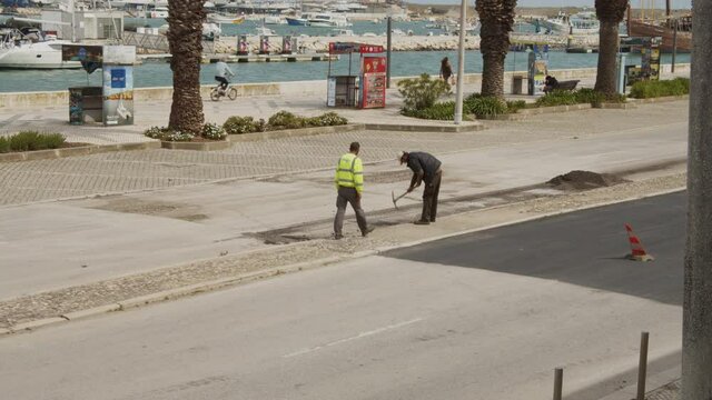 Two men at work. Digging damaged asphalt surface with pickaxe. Local road repair near city marina. Street in Lagos, Portugal
