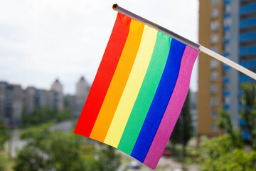 Rainbow LGBT flag set against blue sky, green trees, city street and multi-storey buildings. The concept of tolerance for same-sex couples. Equality for lesbian, gay, bisexual and transgender etc.