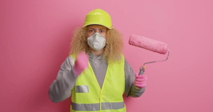 Displeased irritated curly woman repairer clenches fist from anger holds painting roller expresses negative emotions wears hardhat protective respirator and uniform isolated over pink background
