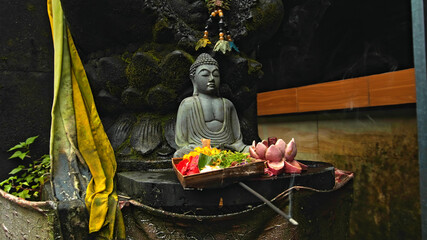 An old stone Budist statue sitting in a lotus pose covered with moss with a wreath of yellow flowers on its neck standing on the street