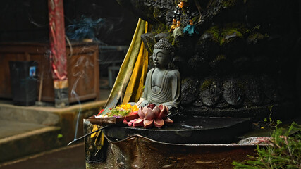 An old stone Budist statue sitting in a lotus pose covered with moss with a wreath of yellow...
