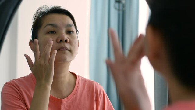 Middle aged asian woman applying cream to her face in the mirror, woman caring of her skin and wrinkles on the face. skin care concept.
