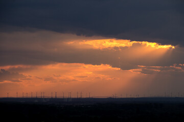 A wide landscape with many wind turbines in warm orange and red evening sunlight shining in rays through a blanket of clouds