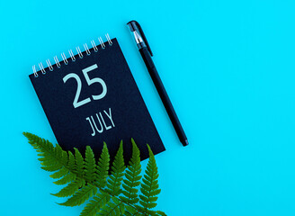 July 25th. Day 25 of month, Calendar date. Black notepad sheet, pen, fern twig, on a blue...