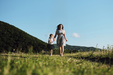 Have fun with child in nature, the joy of motherhood. Young beautiful European mother in blue dress runs through chamomile field with her little daughter and laughs. Front view.